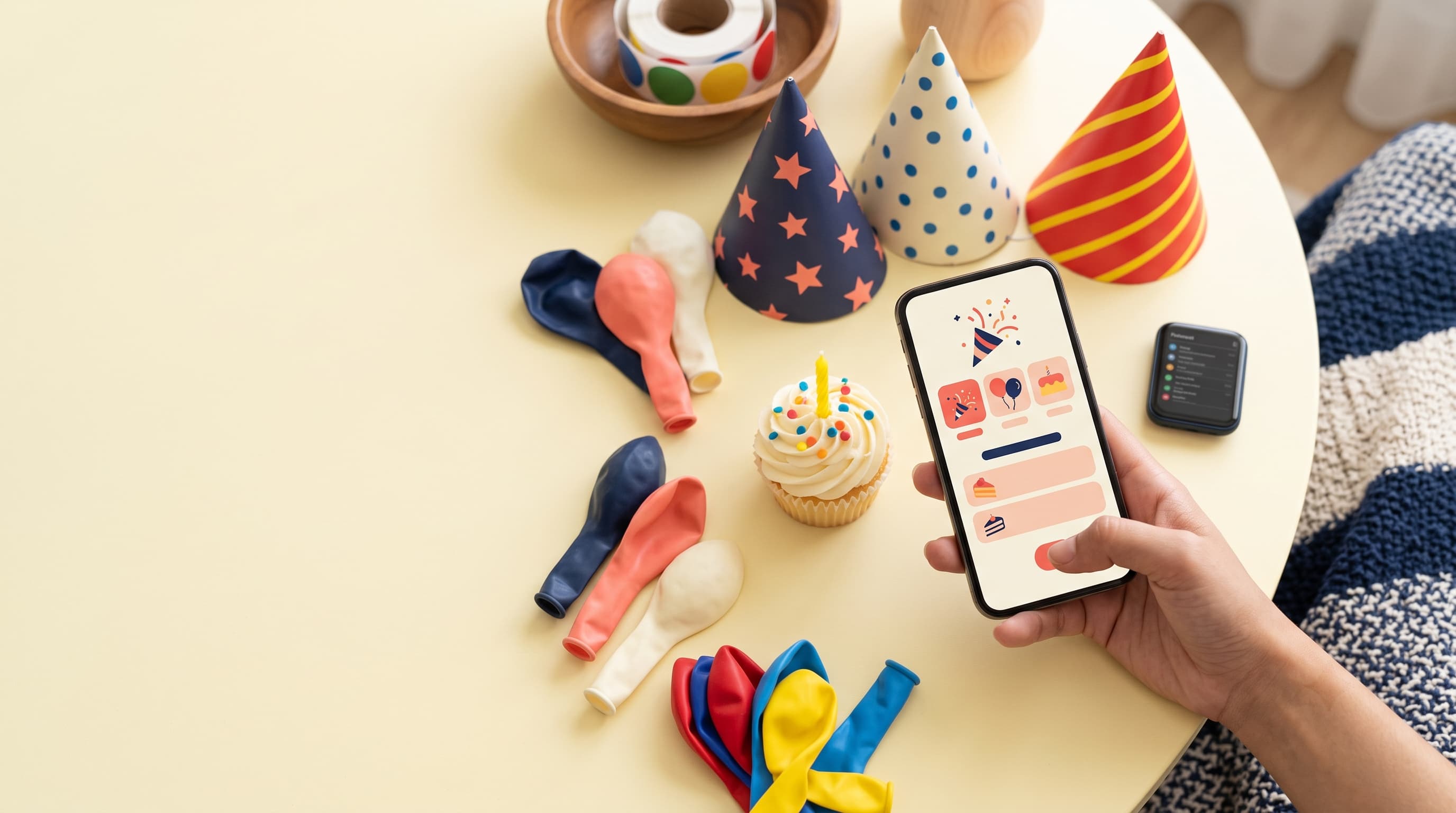 Balloons, party hats, a cupcake and a phone showing a party RSVP screen on a yellow table